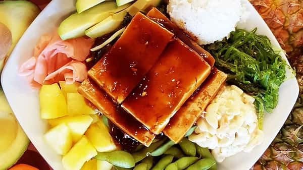 Hawaiian plate lunch with glazed teriyaki tofu slices, avocado, seaweed salad, rice, macaroni salad, pickled ginger, edamame, and pineapple – a flavorful vegetarian island dish.