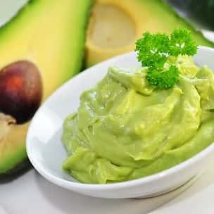 Smooth and creamy guacamole served in a white bowl, garnished with a parsley sprig, alongside fresh avocado halves – a vibrant, healthy dip or topping.