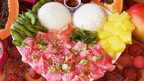 Generous portion of ahi tuna poke garnished with scallions and sesame seeds, served with twin scoops of white rice, seaweed salad, pineapple, edamame, and pickled ginger – a vibrant and fresh Hawaiian poke platter.
