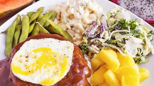 Traditional Hawaiian loco moco featuring a hamburger patty over rice, topped with a sunny-side-up egg and brown gravy, served with edamame, macaroni salad, pineapple, and cabbage slaw.