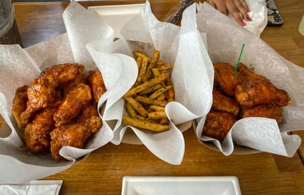 Crispy seasoned wings in two baskets with a center basket of garlic herb fries, all served on parchment paper over a wooden table.