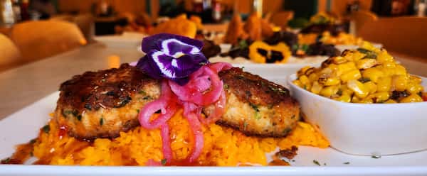 Seared salmon cakes on a bed of seasoned yellow rice, topped with pickled red onions and a purple pansy, served with a side of roasted corn.