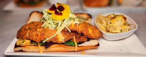 Crispy fried fish fillet on a toasted hoagie roll with slaw and a drizzle of sauce, served with a side of creamy potato salad and an edible flower.