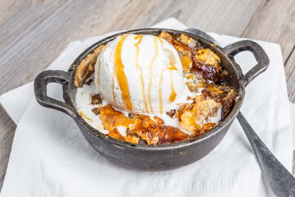 Apple cobbler topped with a scoop of vanilla ice cream and caramel drizzle, served in a cast iron skillet, at a restaurant