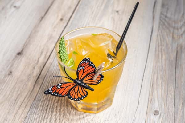 Close-up of a Monarch Smash cocktail, an orange colored drink garnished with mint and a straw, decorated with a monarch butterfly, in a glass on a wooden table at a restaurant