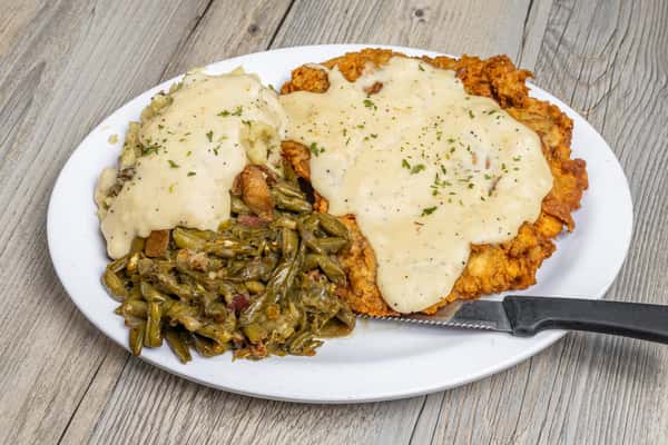 Chicken fried steak topped with white cream gravy, served with a side of sautéed green beans on a white plate, on a wooden table at a restaurant