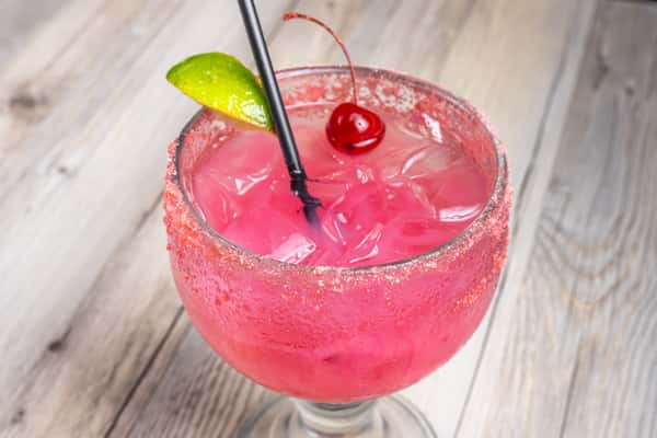 Close-up of a pink Prickly Pear Margarita with a salted rim, garnished with a lime wedge and cherry, in a glass on a wooden table at a restaurant