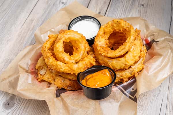 Close-up of golden fried onion rings served with two dipping sauces in a paper-lined basket, on a wooden table at a restaurant
