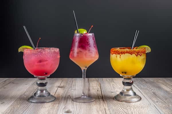 Three colorful cocktails lined up, including a pink margarita and an orange margarita with salted rims and a red layered drink with fruit garnish in the middle, on a wooden table at a restaurant