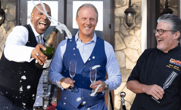 Three smiling men celebrate outside The Winery Restaurant. One man joyfully pops a bottle of champagne, with foam spraying into the air. The man in the center holds two empty champagne flutes and smiles at the camera, while the chef on the right, wearing a black uniform with The Winery logo, laughs and holds a glass. The background shows stone walls and elegant lanterns by the entrance.