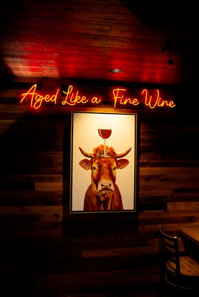 Neon sign reading "Aged Like a Fine Wine" above artwork of a cow with a glass of red wine balanced on its head.