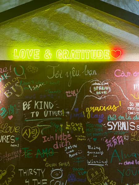 Wall with neon sign reading "LOVE & GRATITUDE ❤️" above colorful handwritten messages and doodles in multiple languages.