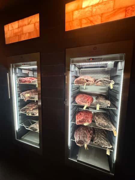 Illuminated dry-aging refrigerators displaying large cuts of beef on metal racks in a dimly lit restaurant setting.
