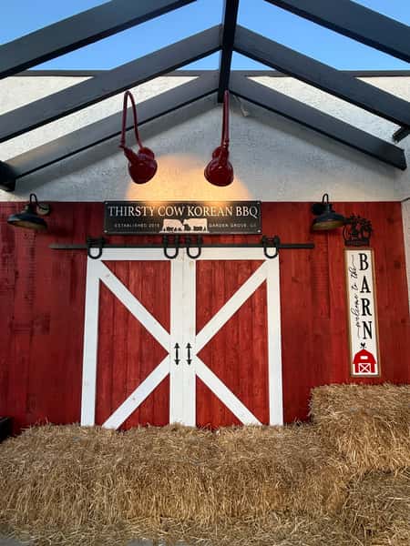 Barn-themed exterior at Thirsty Cow Korean BBQ with red sliding doors, hay bales, and rustic lighting fixtures.