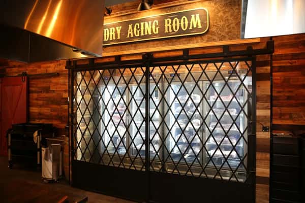 Restaurant dry aging room with refrigerators storing cuts of meat, viewed through black lattice doors under a wooden sign.