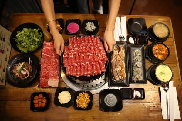 A Korean BBQ feast with assorted raw meats, side dishes, soups, and vegetables arranged neatly on a wooden table.