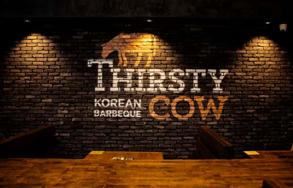 Thirsty Cow Korean Barbeque logo painted on a brick wall, lit by warm spotlights above wooden dining booths.