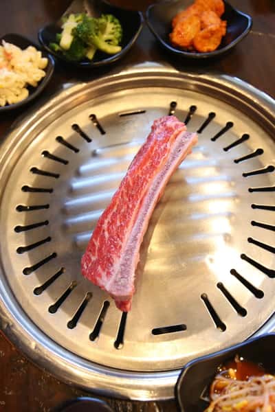 Raw beef short rib placed on a Korean BBQ tabletop grill, surrounded by side dishes like kimchi, broccoli, and macaroni salad.