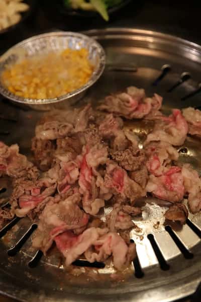 Thin slices of beef grilling on a Korean BBQ tabletop grill with a side of cheesy corn in a foil dish.