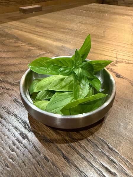 Small metal bowl filled with fresh green basil leaves placed on a wooden table.