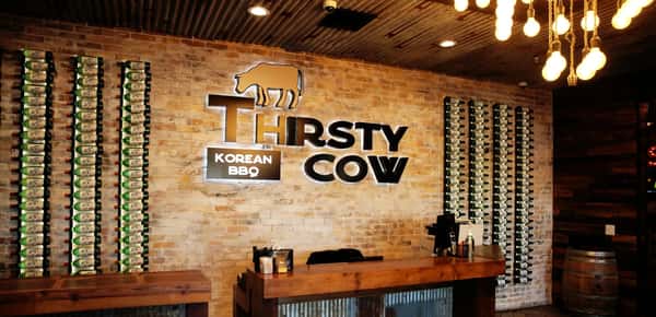 Thirsty Cow Korean BBQ restaurant reception area with illuminated logo, brick wall, wooden counters, and soju bottle displays.