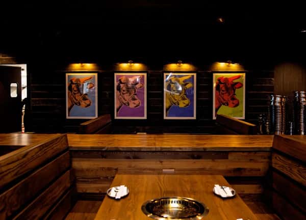 Restaurant interior with wooden booths and grill tables, featuring colorful cow artwork displayed on the dark wood-paneled wall.