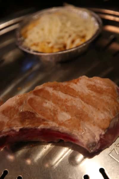Thick cut of meat grilling on a Korean BBQ tabletop grill with a side dish of cheesy corn in foil nearby.