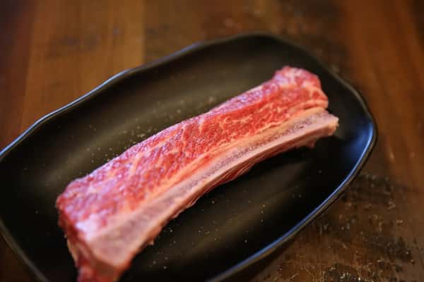 Raw beef short rib placed on a Korean BBQ tabletop grill, surrounded by side dishes like kimchi, broccoli, and macaroni salad.