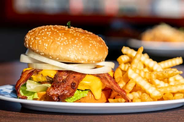 A stacked cheeseburger served on a sesame seed bun with melted cheese, fresh toppings, and a juicy beef patty, accompanied by a side of crispy French fries. The close-up angle highlights the rich textures and classic comfort-food appeal.