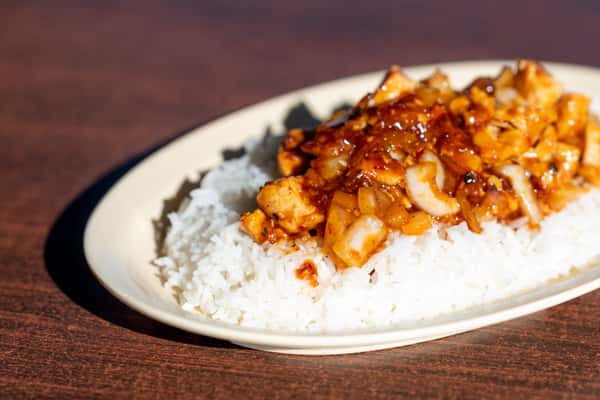 A plate of white rice topped with diced grilled chicken glazed in a glossy savory sauce and sprinkled with sesame seeds. The dish has a warm, homemade feel with a balance of sweet and savory flavors.