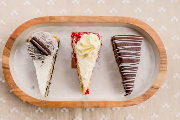 Three slices of cheesecake including chocolate, strawberry, and classic varieties on a wooden board from The Butcher Shop bakery