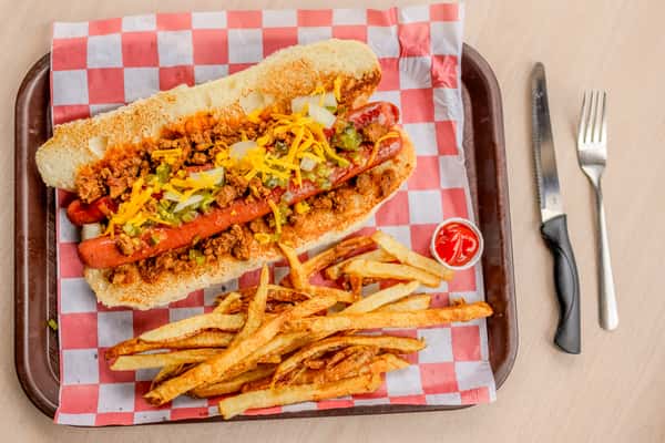 A footlong hot dog topped with chili and cheese served with seasoned fries on a checkered tray at The Butcher Shop restaurant