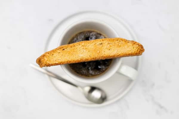 Overhead shot shows a biscotti resting on a mug of coffee. The mug and biscotti sit on a white plate, with a spoon underneath.