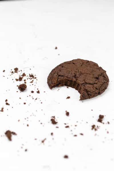 A partial chocolate cookie sits on a white surface, surrounded by crumbs. The bite mark suggests that the cookie is likely delicious and freshly made.