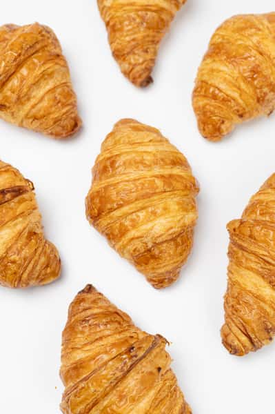 Seven golden brown croissants are arranged on a white surface. The pastries appear freshly baked, showcasing their flaky layers and glossy finish.