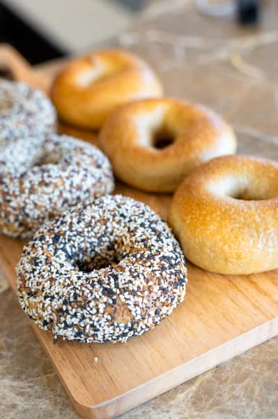 A wooden cutting board with bagels. Some bagel are covered in white and black seeds, and others are plain.