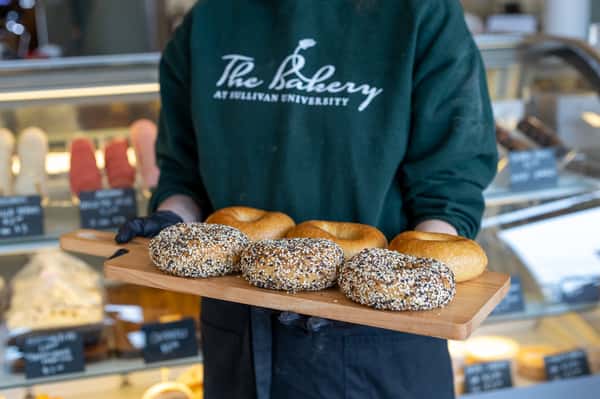 A person holds a wooden tray of bagels, three with everything-seasoning. The person wears an apron and a sweatshirt that reads: "The Bakery at Sullivan University".