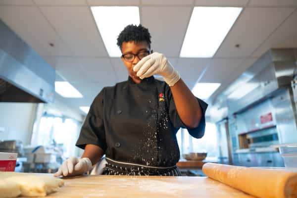 A pastry chef in a black uniform is sprinkling flour on a wooden counter, preparing dough. The image promotes The Broidery at Southern University's website.
