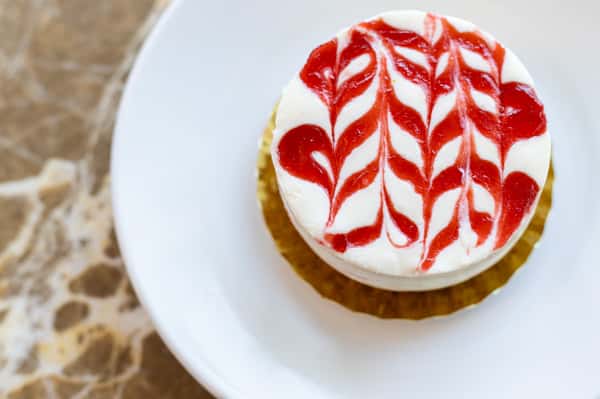 Here is a top-down shot of a cheesecake with an intricate swirl of red jam on top. It sits on a white plate, ready to be described for visually impaired customers.