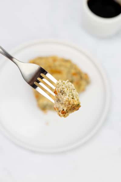 Here's a description of the image, perfect for alt text: A piece of baked savory cake is held on a fork above a white plate. The cake has flecks of green and orange, and a cup is out of focus in the background.