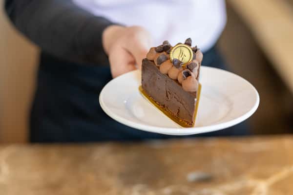 A server holds a slice of chocolate cake topped with chocolate chips and a gold emblem. The cake is on a white plate, presented on a counter for a restaurant or bakery.