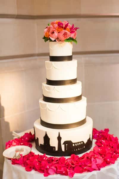 Five-tier white wedding cake with black ribbon accents, topped with pink flowers, surrounded by red rose petals on a table.