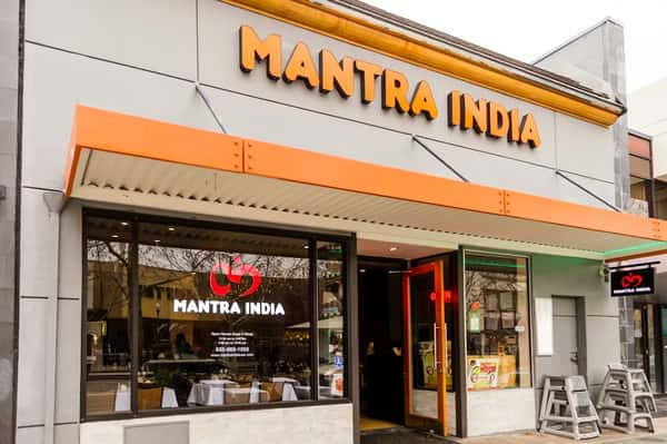 Front exterior of Mantra India restaurant with orange awning, glass windows, and an open door inviting customers inside.