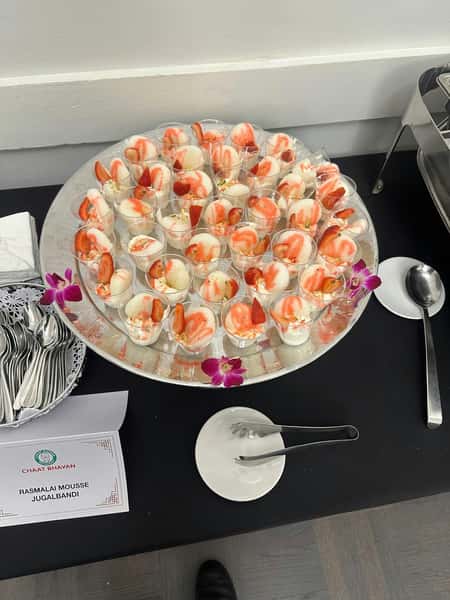 Tray of Rasmalai Mousse Jugalbandi cups garnished with strawberry slices and drizzled syrup, surrounded by orchid flowers.