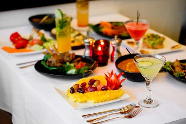 Elegant dining setup with assorted Indian dishes, cocktails, and a decorative fruit plate with sliced mango, grapes, and carved strawberries.