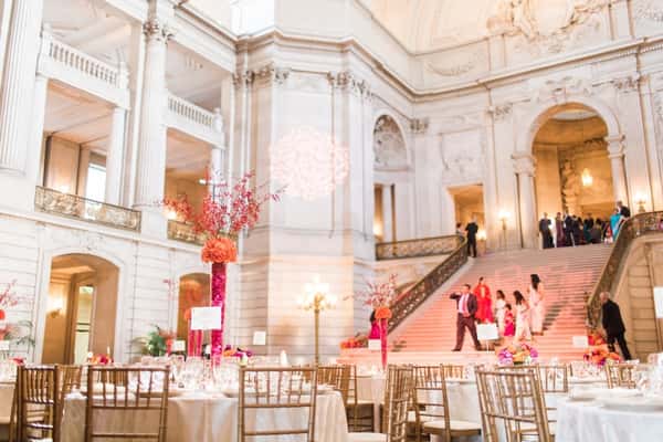 Elegant wedding reception setup in a grand hall with high ceilings, floral centerpieces, and guests ascending a grand staircase.