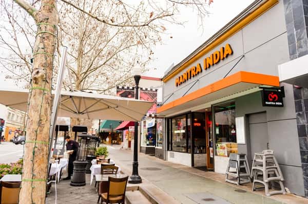 Street view of Mantra India restaurant with outdoor dining tables under large umbrellas on a tree-lined sidewalk.