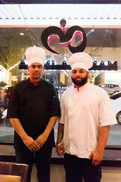 Two chefs wearing traditional white hats stand in front of a window with a restaurant logo, dressed in black and white uniforms.