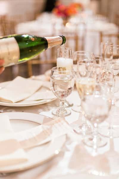 Champagne being poured into a flute glass at a formal event table set with elegant plates, napkins, and multiple types of stemware.