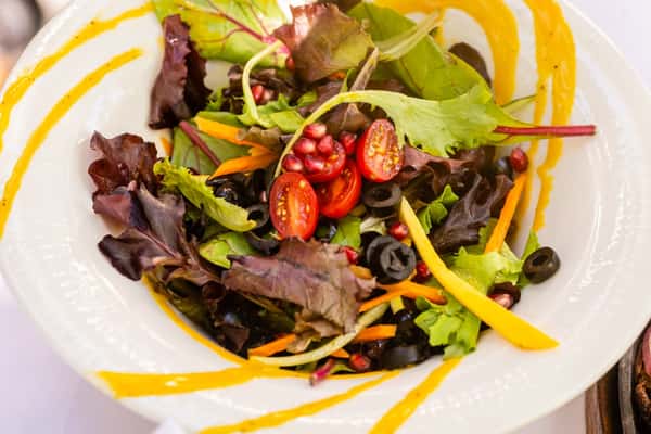 Fresh salad with cherry tomatoes, black olives, pomegranate seeds, and mixed greens garnished with yellow dressing swirls.
